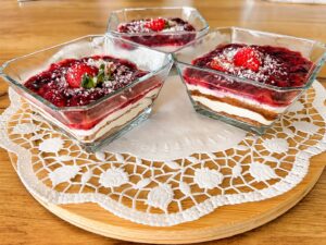 Four square glass bowls on a lace doily each with layered berry sauce cream and biscuit crumbs on a wooden table